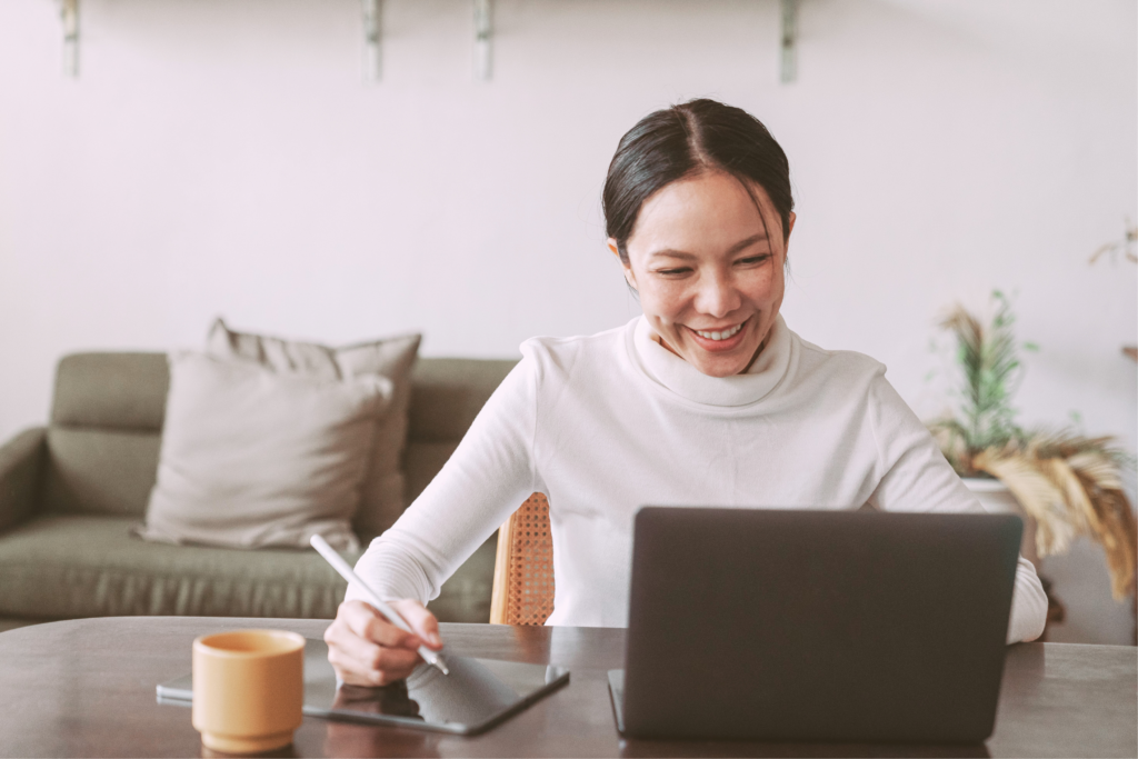 Asian female virtual assistant smiling at the computer while writing notes and seemingly being in an online meeting 